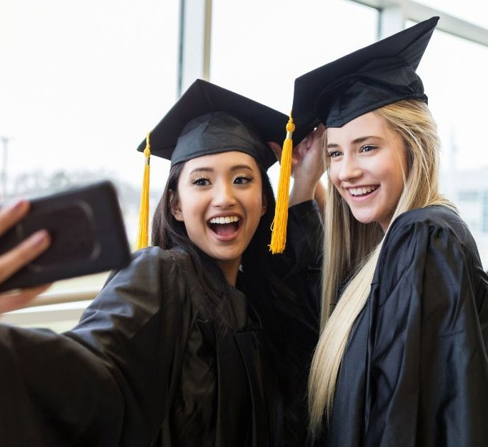 grads2 two girls in black caps and graduation gowns taking a selfie